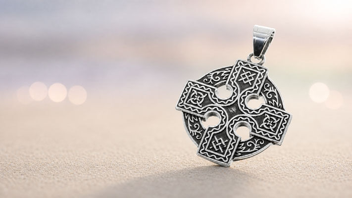 Silberfarbener runder Keltenkreuz-Anhänger mit detailreichen Ornamenten vor unscharfer Küstenkulisse.