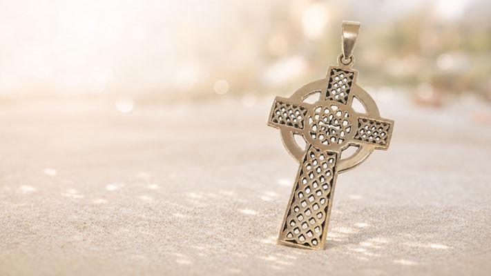 Goldfarbener Keltenkreuz-Anhänger mit filigranem Gittermuster auf hellem Sand.